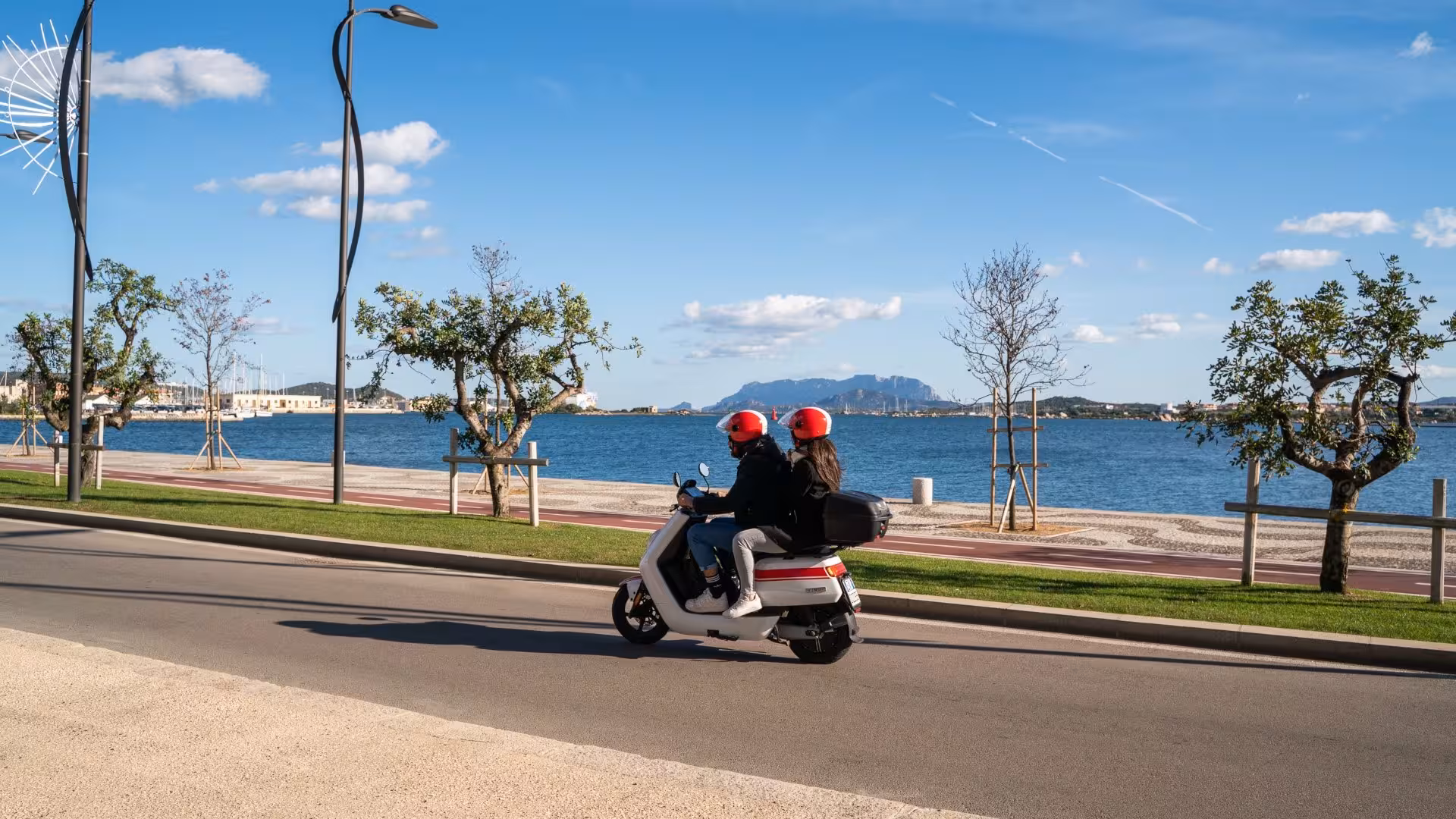 Two people riding a scooter along Olbia's beautiful waterfront, showcasing a perfect rental adventure.