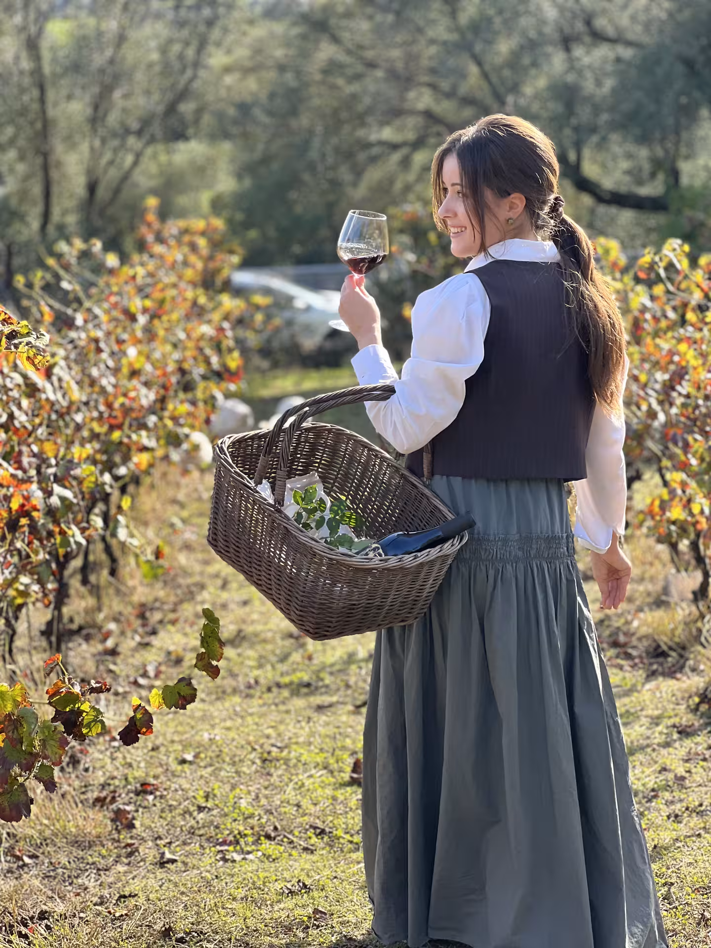Woman enjoying a vineyard picnic in Olbia, holding a wine glass and basket amidst lush grapevines.