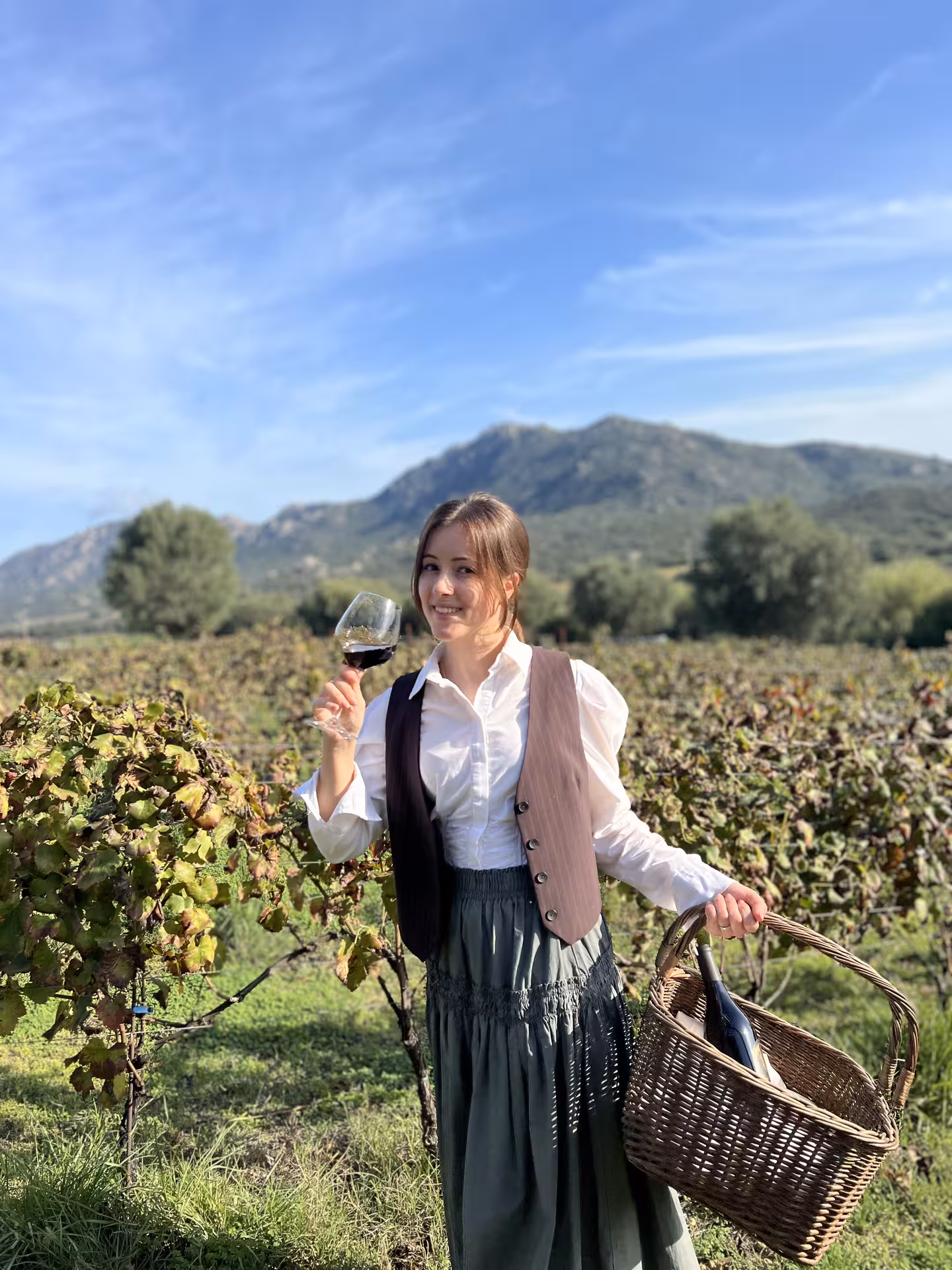 Smiling woman toasting with red wine in an Olbia vineyard, surrounded by scenic hills and grapevines.
