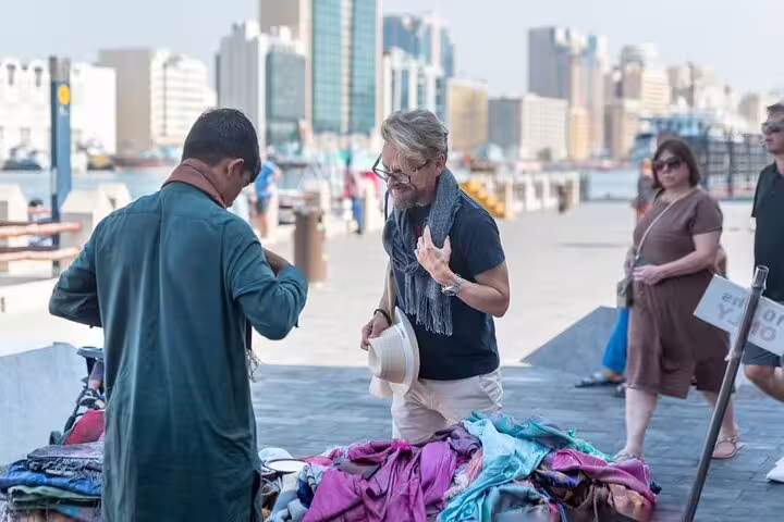 Tourists browse colorful scarves at a waterfront market stall during a guided walking tour of old Dubai with local snacks