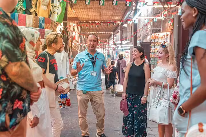 Small group on guided walking tour of Dubai Old Town, listening to local guide in colorful souk lined with snacks and souvenirs