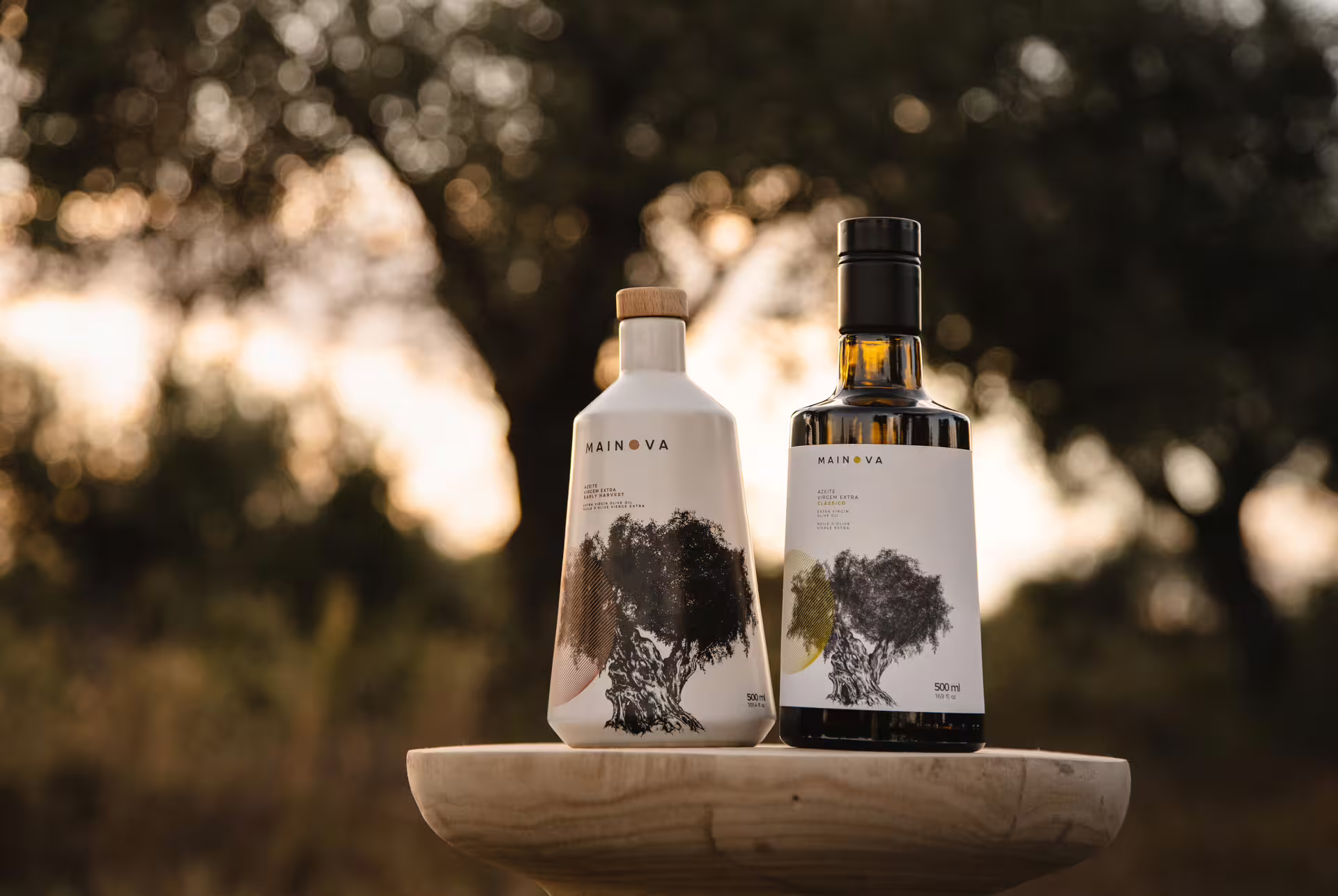 Two elegantly labeled olive oil bottles on a wooden stand, set against a blurred background of olive trees at sunset.