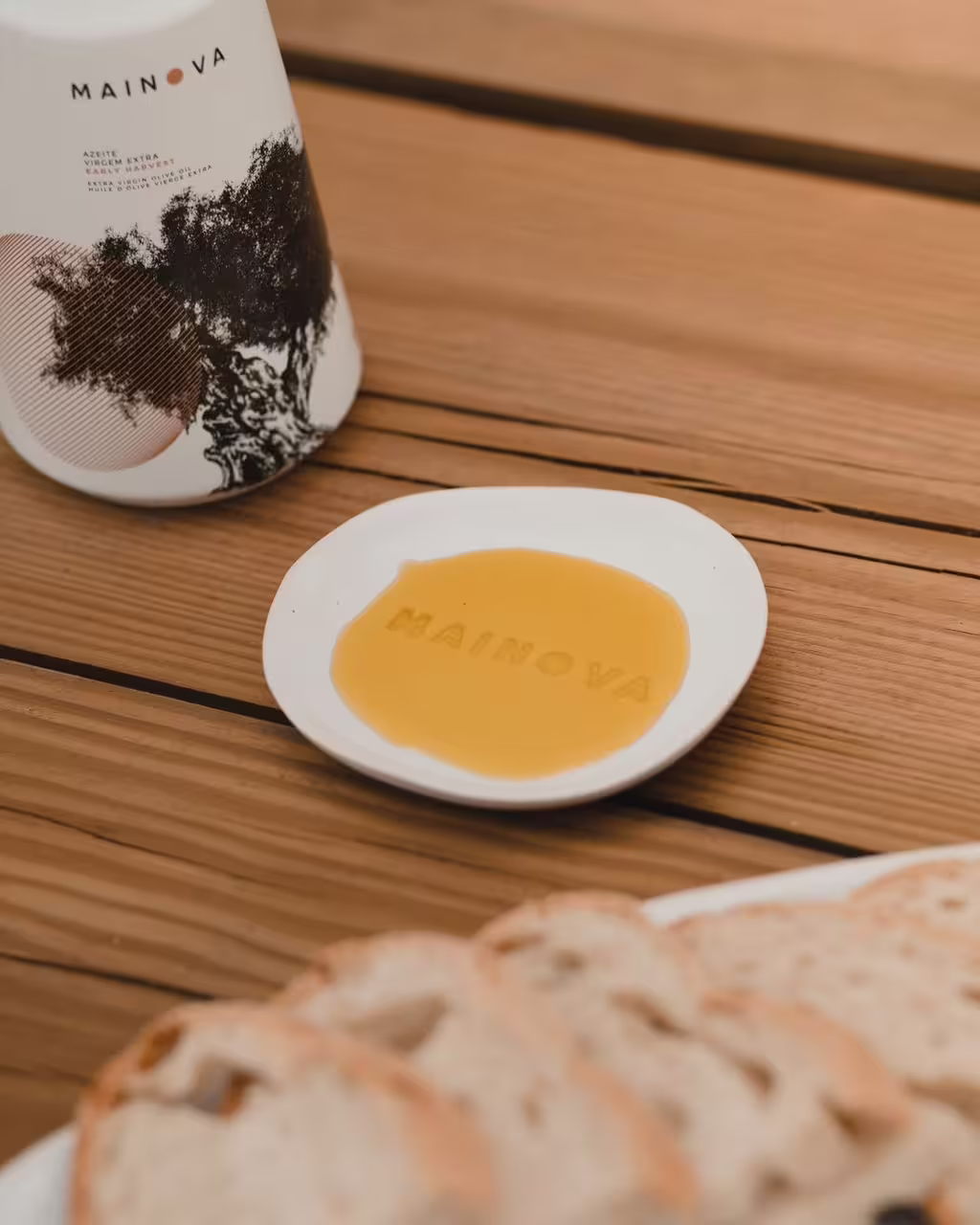 Plate with olive oil labeled "Mainova" next to a bottle and sliced bread on a wooden table, highlighting tasting experience.