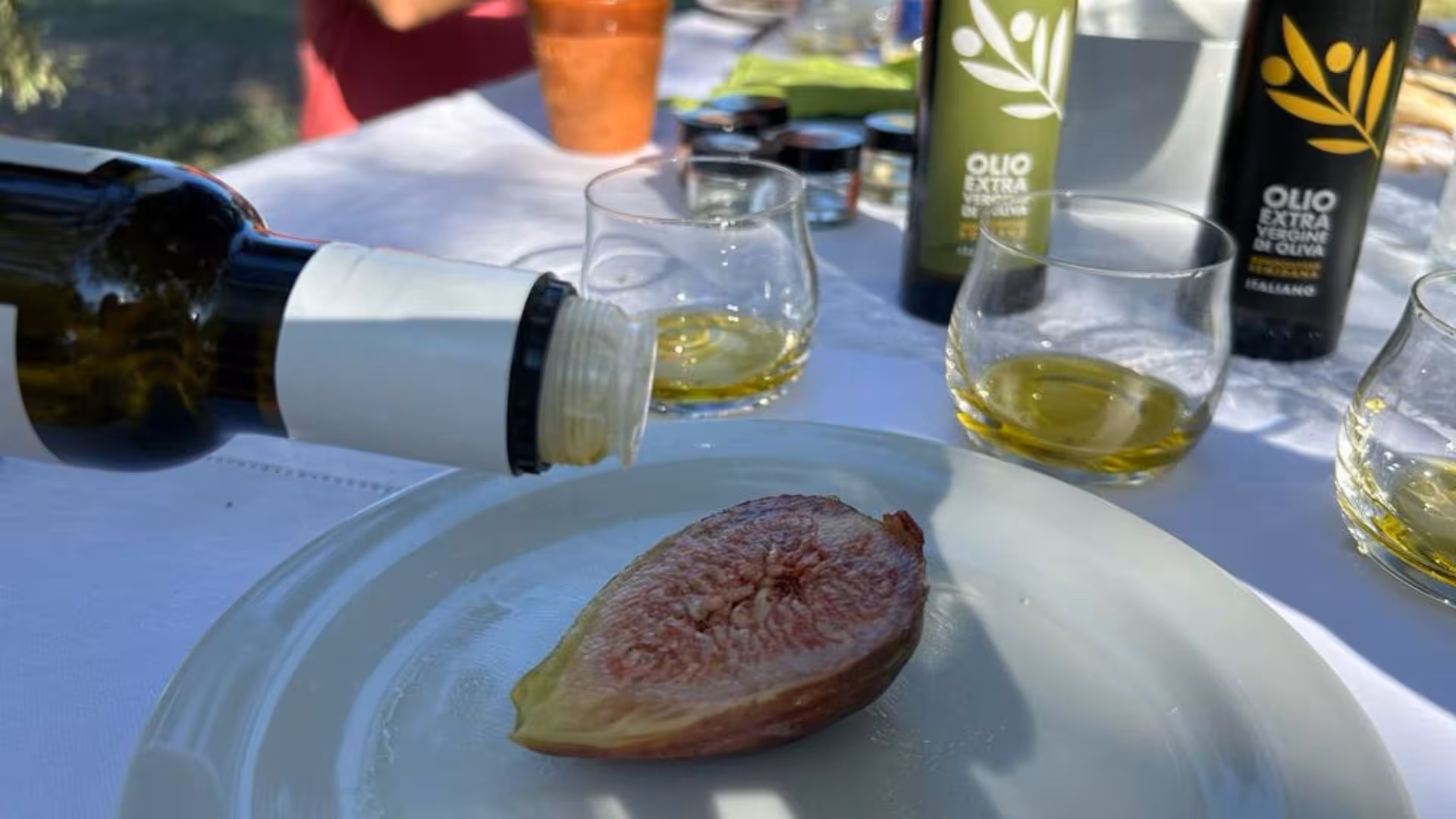 Pouring olive oil over fresh fig at a tasting table in Oristano, surrounded by glasses and olive oil bottles.