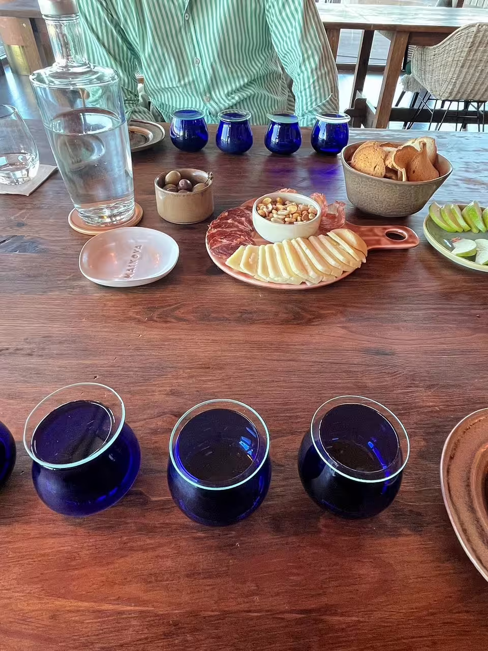 Olive oil tasting setup with six blue glasses, regional cheeses, cured meats, olives, nuts, and bread on a wooden table.