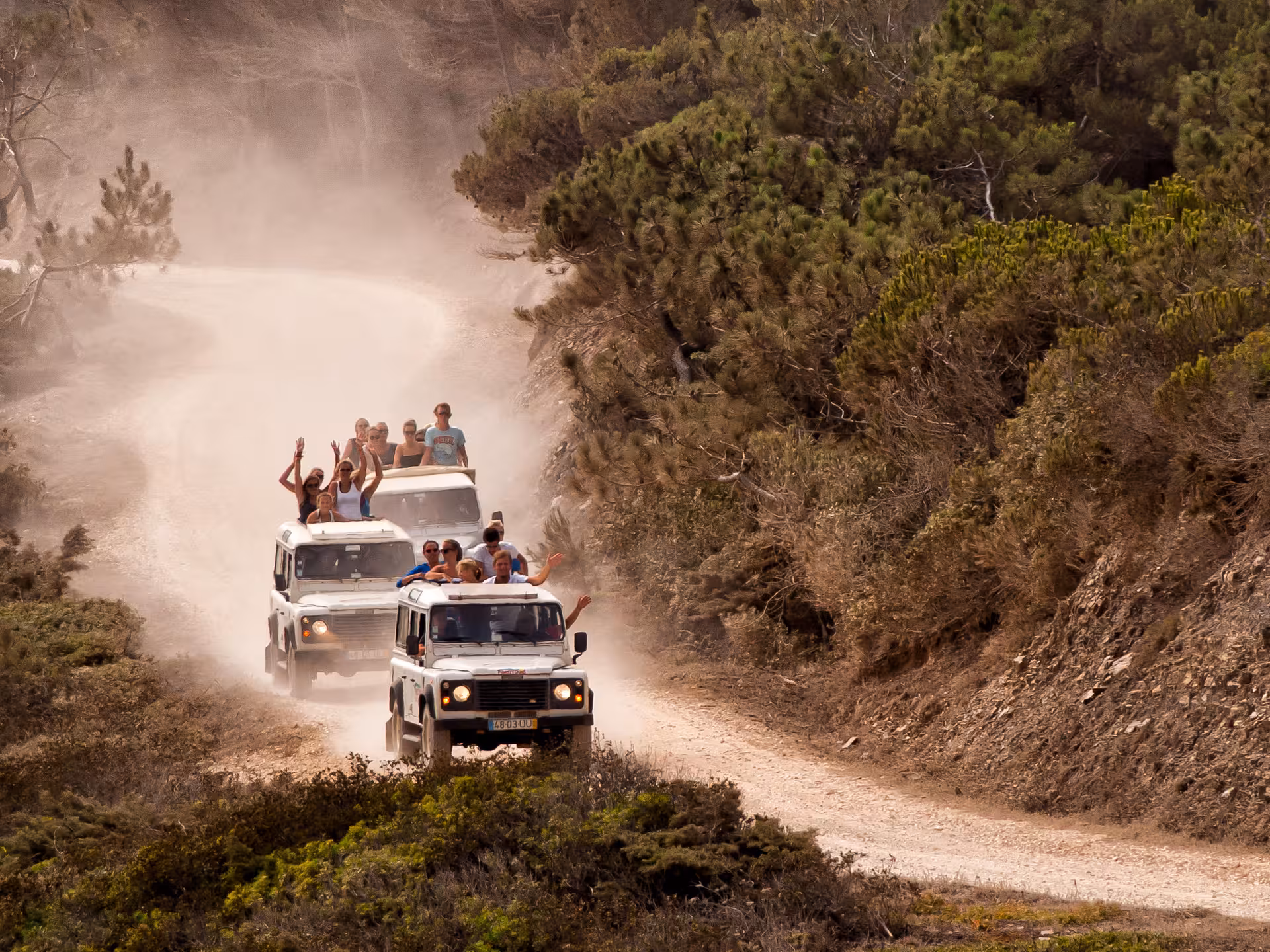 Open-top Jeeps full of smiling travelers drive a dusty forest trail on an Algarve off-road Jeep safari adventure tour