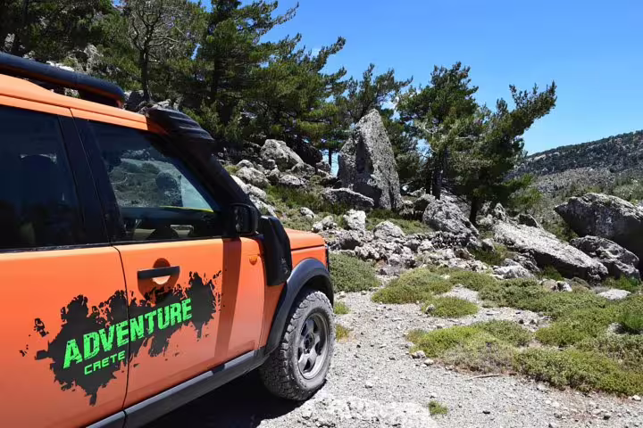 Orange adventure vehicle on rocky terrain in Crete's Lassithi Plateau, surrounded by lush greenery and clear blue skies.