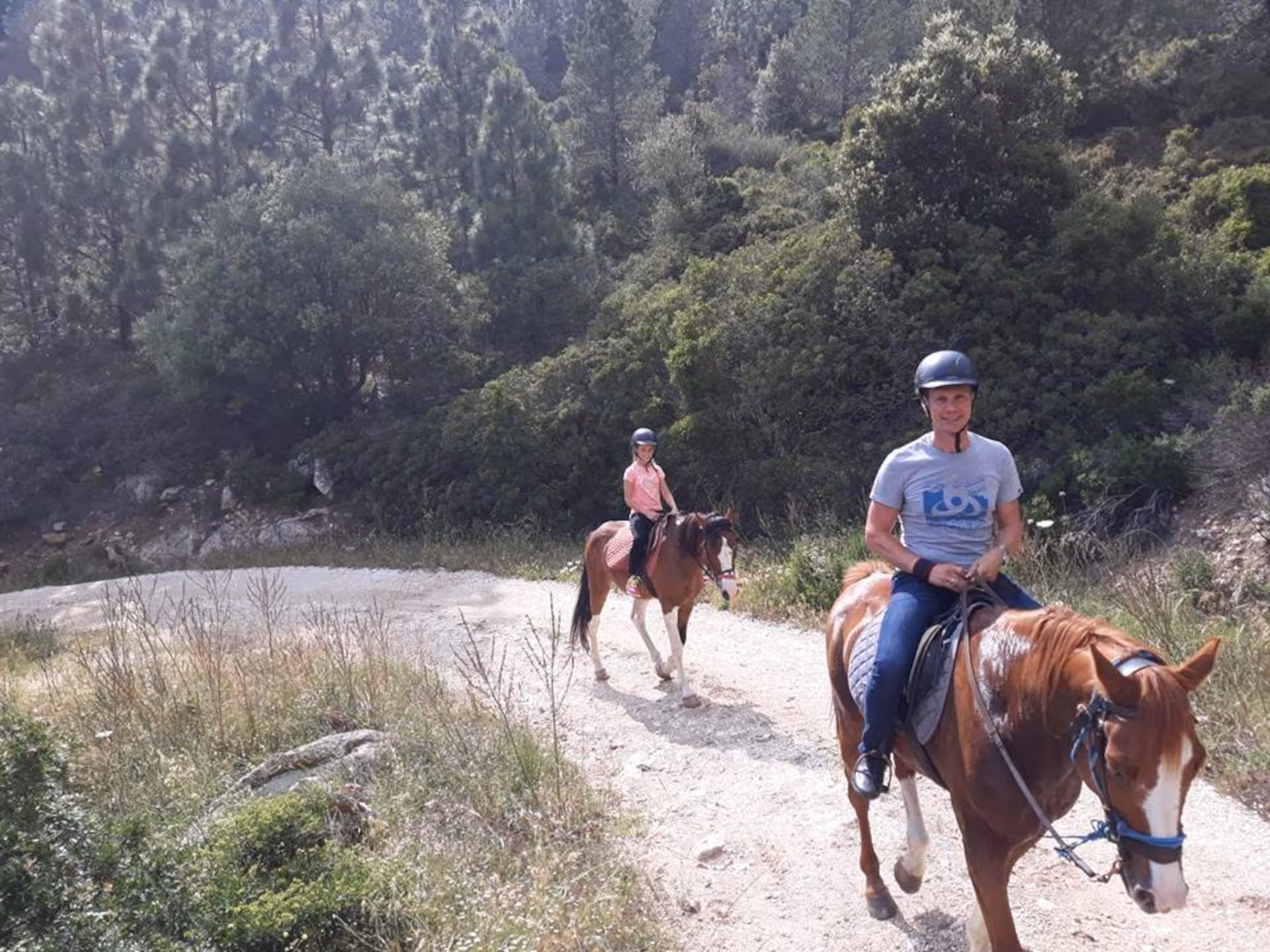 Two riders horseback riding through a sunlit forest trail in Orosei.
