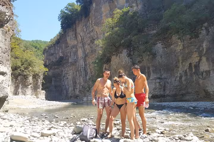 A group of friends explores the rocky riverbank of Osum Canyon, surrounded by towering cliffs and clear waters.