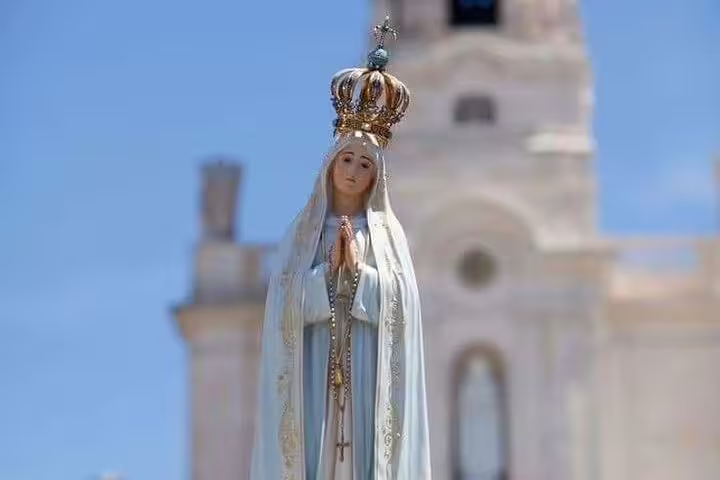 A statue of Our Lady of Fátima with a crown, set against the backdrop of the Sanctuary of Fátima under a clear blue sky.