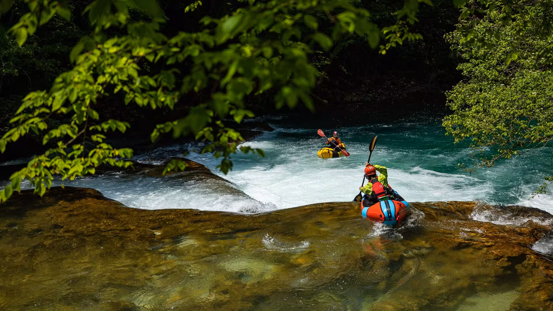 Two packrafters navigating the rapids of Mreznica River surrounded by vibrant green foliage.