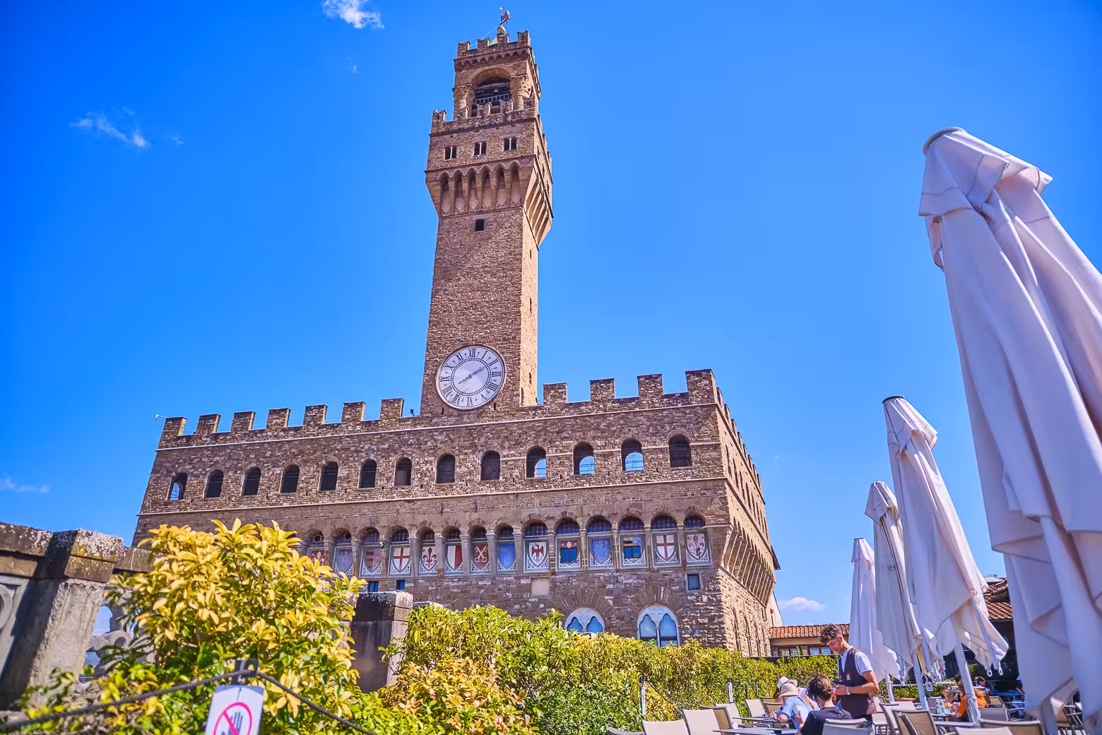 View of the historic tower of Palazzo Vecchio with a clock, seen from a terrace with umbrellas against a blue sky.