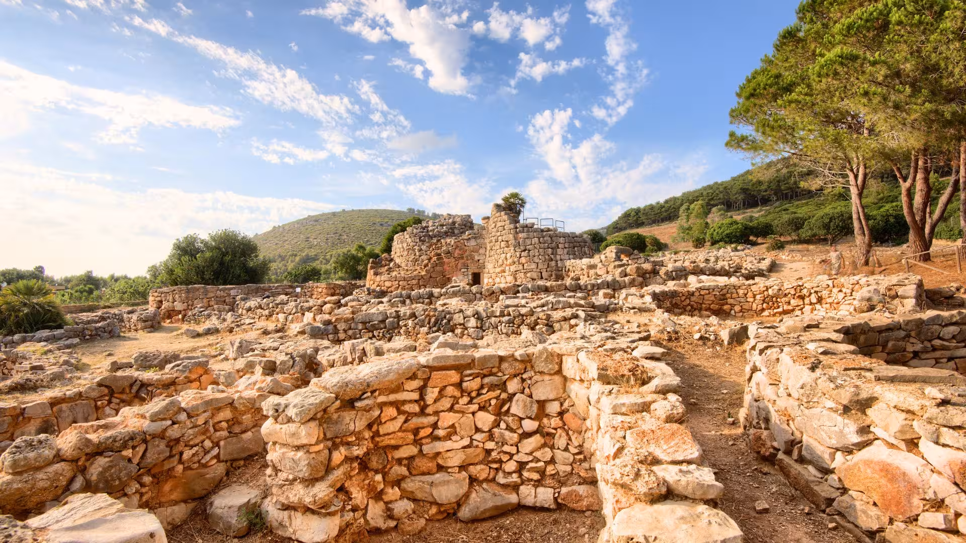 Panoramic view of Palmavera nuraghe ruins in Alghero, with scenic hills and trees under a bright, blue sky.