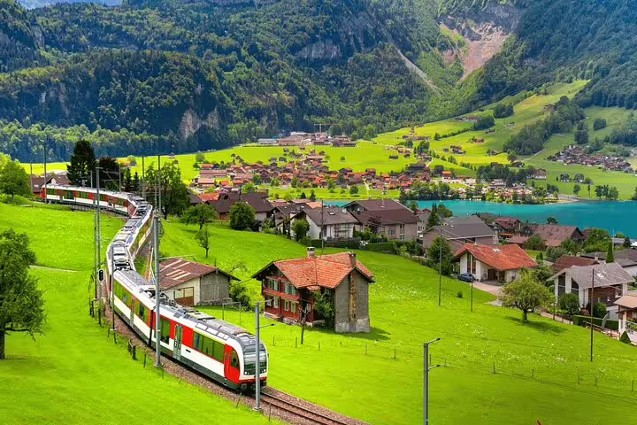Panoramic train traversing picturesque Swiss village, surrounded by vibrant green hills and serene lakeside scenery.