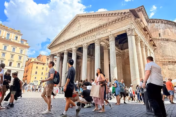 Crowds gather outside the iconic Pantheon in Rome, showcasing its ancient columns and detailed facade under a sunny sky.