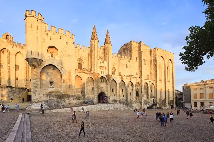 The majestic Papal Palace in Avignon, bathed in golden sunlight, with visitors exploring its historic courtyard.