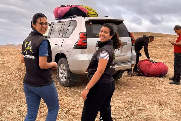 Two women smiling near a 4x4 vehicle with paragliding equipment in Agafay Desert, Marrakech, ready for adventure.