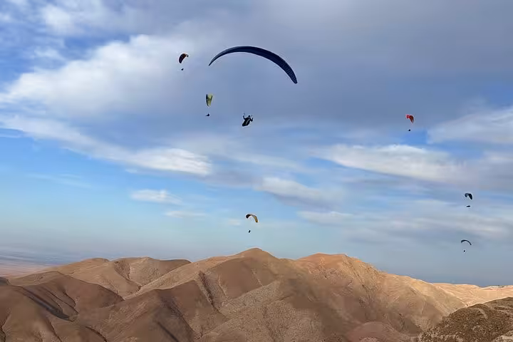 Multiple paragliders glide above the rugged Agafay Desert landscape near Marrakech under a vast, cloudy sky.