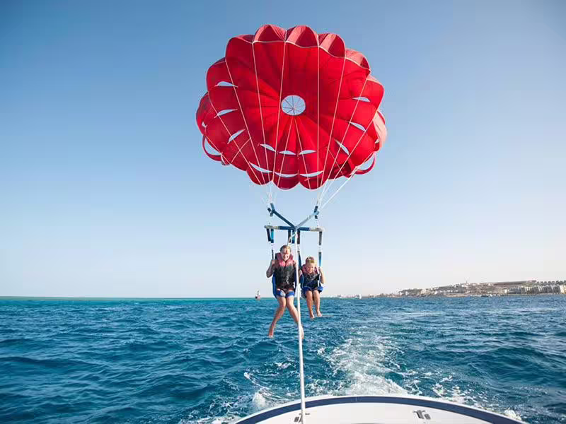 Two riders parasailing behind a boat over the Red Sea in Hurghada, Egypt, with red parachute and coastline views