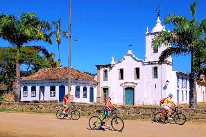 Cyclists explore Paraty’s historic streets, passing a charming white church and colonial buildings under palm trees.