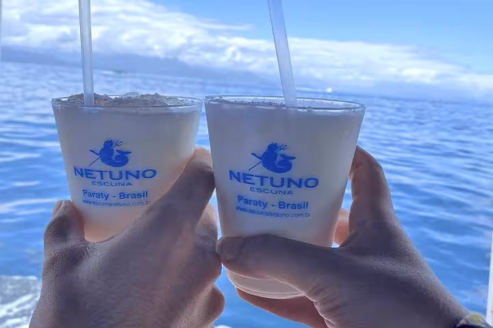 Two hands holding tropical drinks aboard a boat tour in Paraty, Brazil, with scenic ocean views in the background.