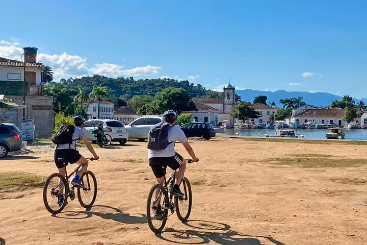 Cyclists riding along the scenic waterfront in Paraty with historic buildings and mountains in the background.