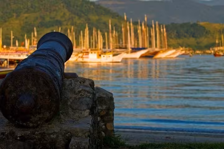 Cannon overlooking the serene bay and sailboats in Paraty, highlighting historical significance on the walking tour.