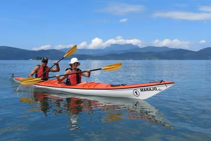 Two people paddling a kayak on calm waters with scenic mountain views during a Paraty beach tour.