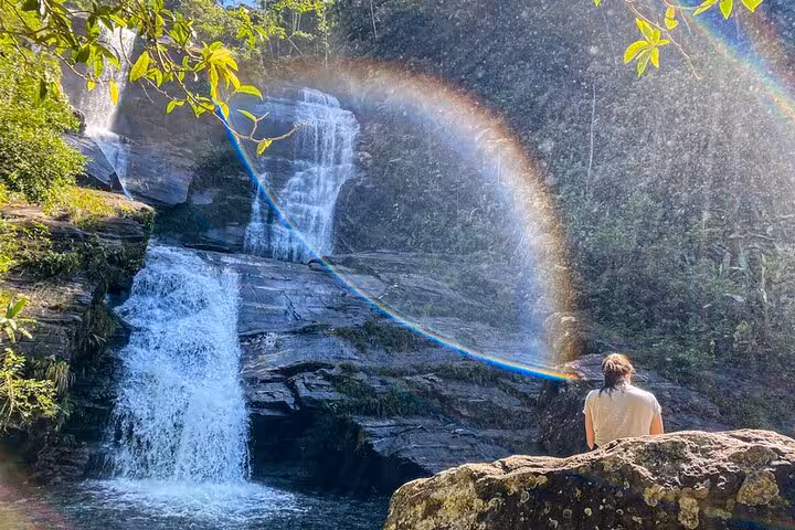 Serene view of a person sitting by Melancia Waterfall with a rainbow, lush greenery, and cascading water in Paraty.