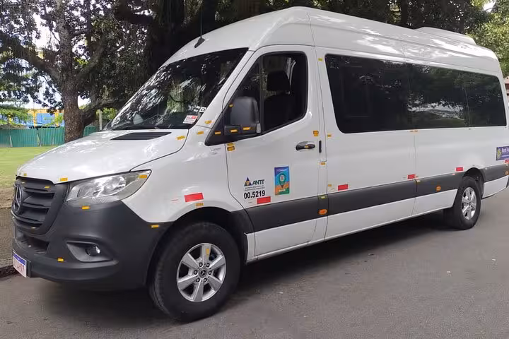 Comfortable white shuttle van ready for shared transfer from Paraty to São Paulo, parked under lush trees.