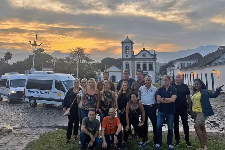 Group of tourists posing in front of Paraty Tours vans with a historic church backdrop during sunset in Paraty.