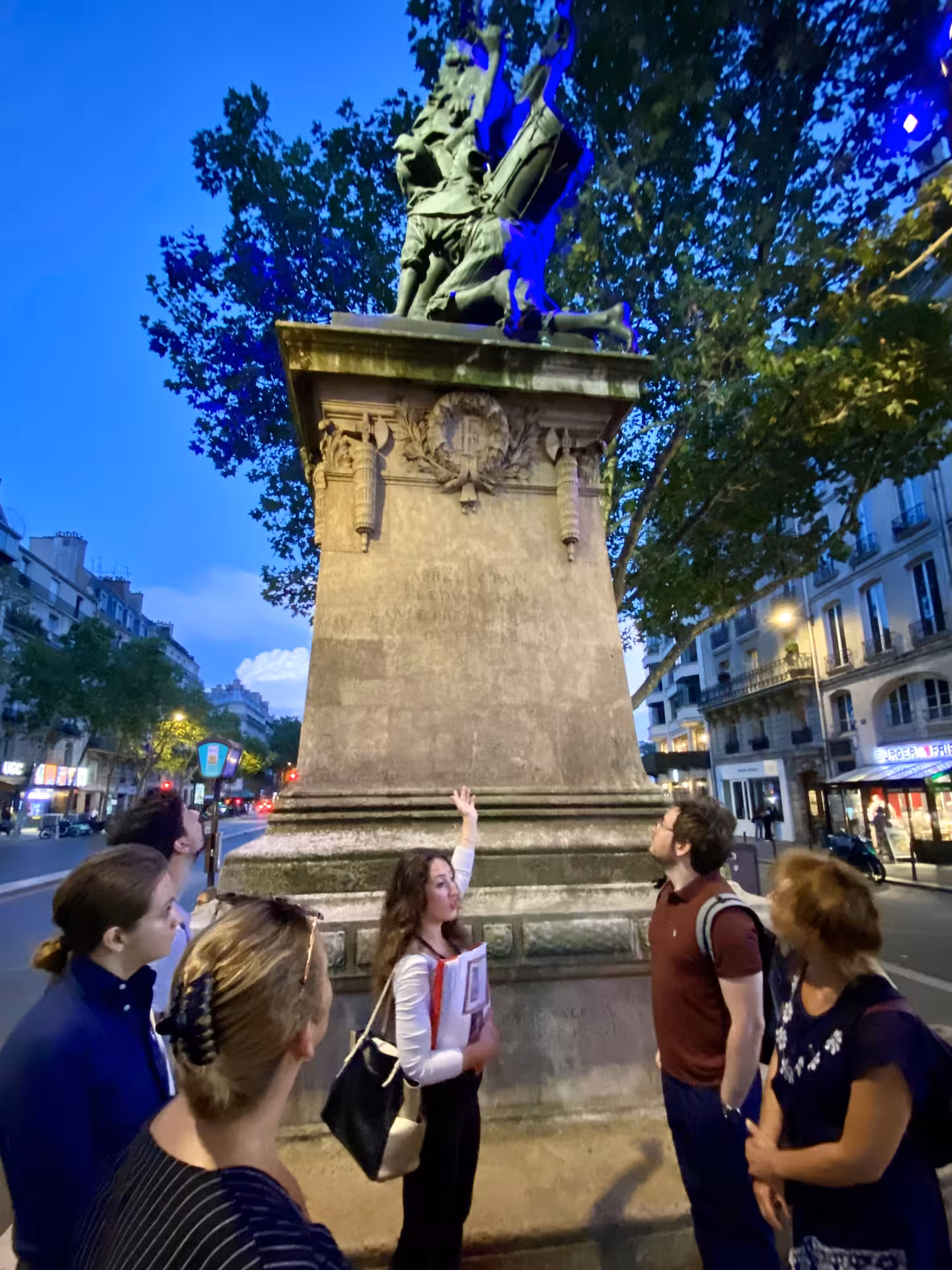 Tour group admires a dramatic French Revolution statue in Paris under evening lights.