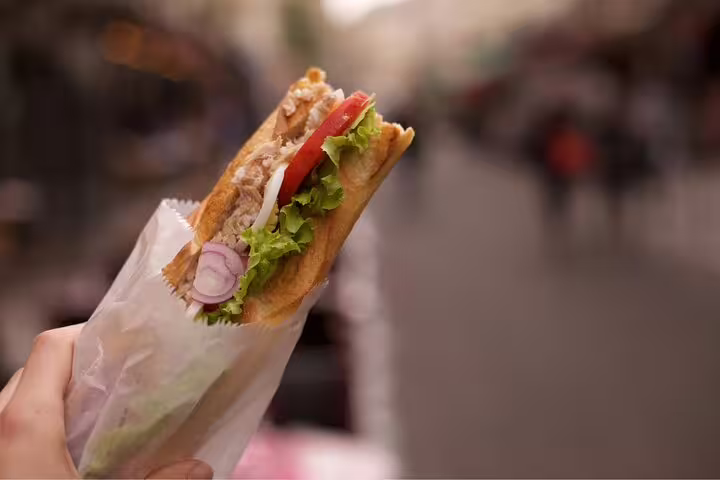 A hand holds a fresh baguette sandwich with lettuce, tomatoes, and tuna on a Paris street during a Le Marais food tour.