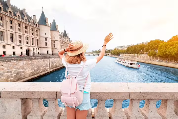 Woman with backpack and sunhat waving from a Paris bridge, overlooking the Seine River with a boat and historic buildings.