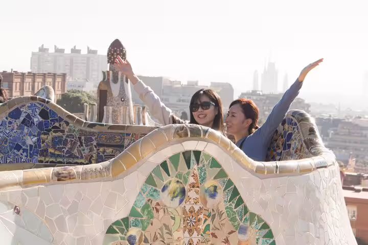 Two people joyfully pose on Park Güell’s mosaic terrace with views of Barcelona cityscape.