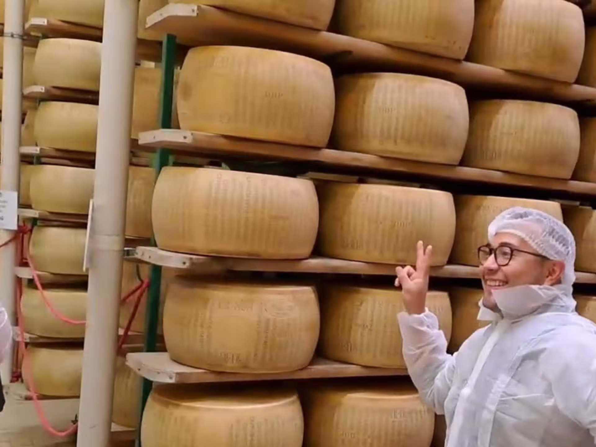 A visitor in protective gear poses with large Parmesan cheese wheels during a factory tour near Parma.