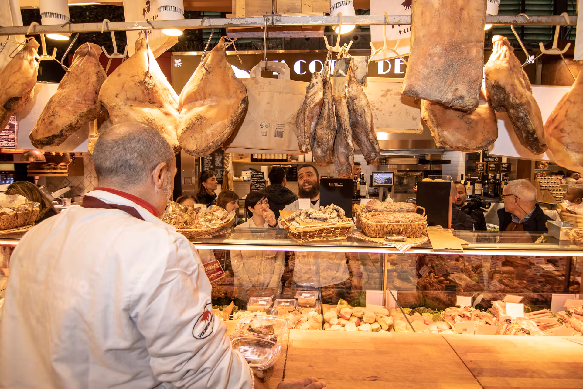 Group admires hanging meats and cheeses at a lively stall in Florence's Sant’Ambrogio Market, part of a culinary tour.