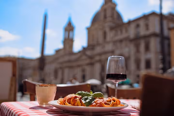 Delicious pasta and a glass of red wine on a checkered tablecloth with Piazza Navona in the background, Rome.