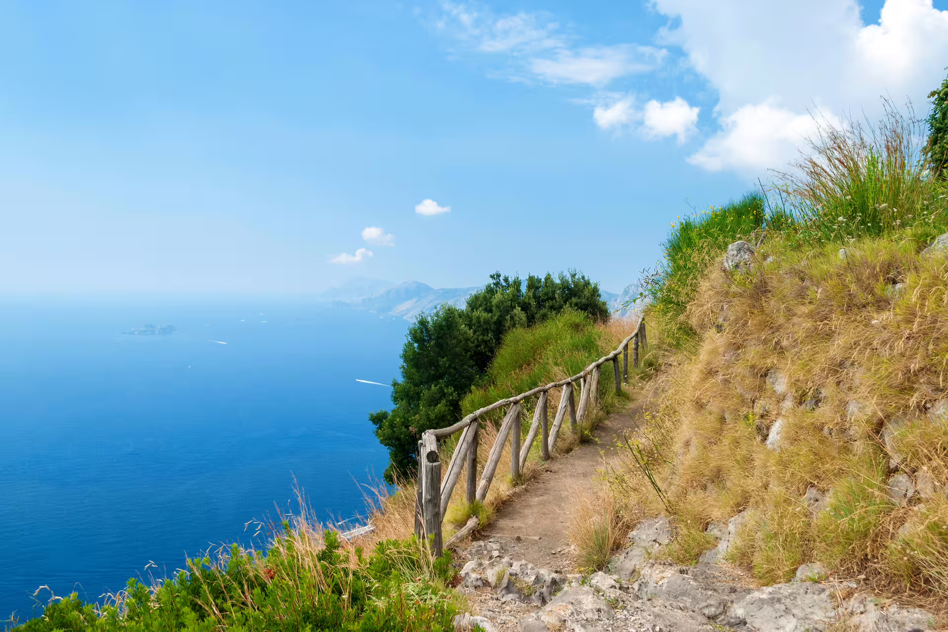 Sunny dirt trail with rustic wooden fence along the Path of the Gods, offering sweeping Amalfi Coast and Capri sea views in Italy