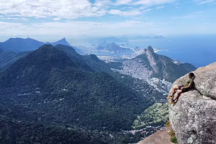 Adventurer relaxing on Pedra da Gávea cliff, overlooking lush forests and iconic Rio de Janeiro cityscape.