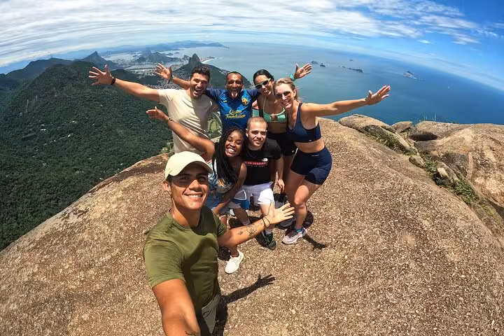Smiling hikers celebrating their ascent on Pedra da Gávea, with panoramic views of Rio and the ocean.