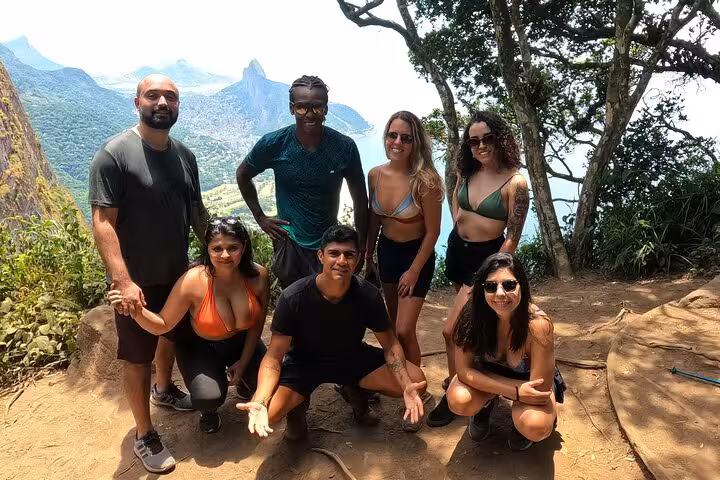 Group of hikers smiling at the summit of Pedra da Gávea, with stunning mountain views in the background.