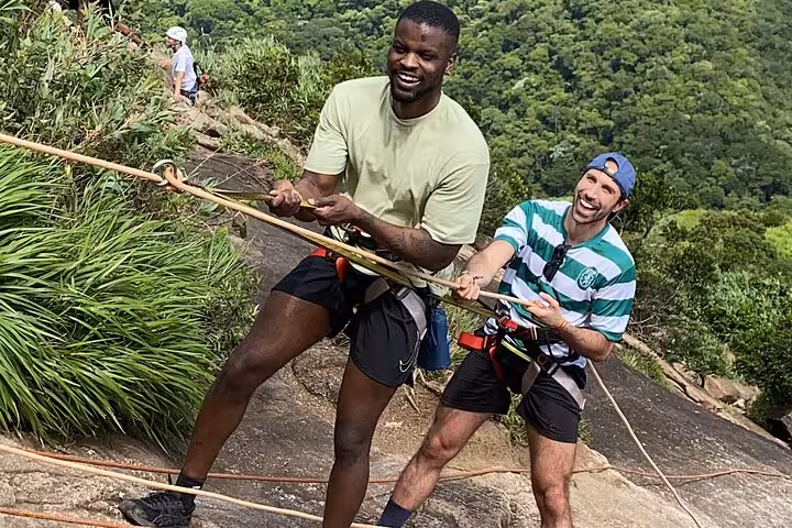 Two hikers enjoy a thrilling climb with ropes on the lush Pedra da Gávea trail in Rio, showcasing adventure and teamwork.