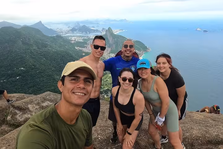 A group of six friends celebrates reaching the peak of Pedra da Gávea, with stunning views of Rio de Janeiro's coastline.