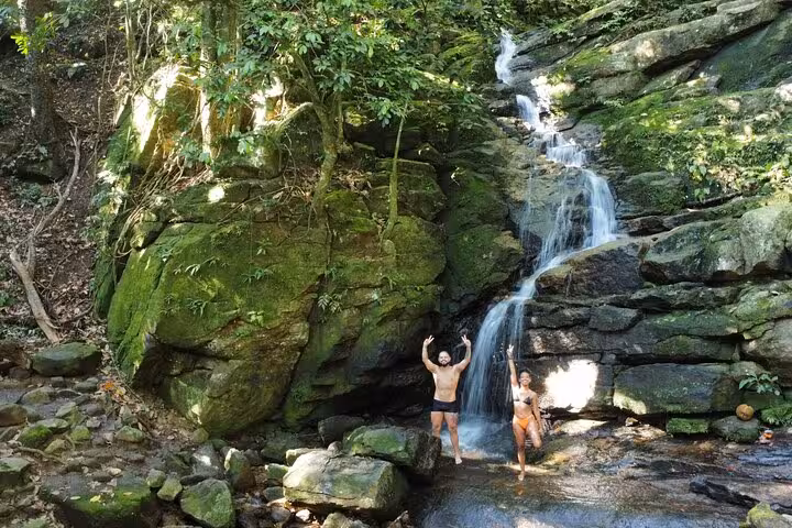 Two adventurers reveling under a serene waterfall surrounded by lush greenery on the Pedra da Gávea hike.