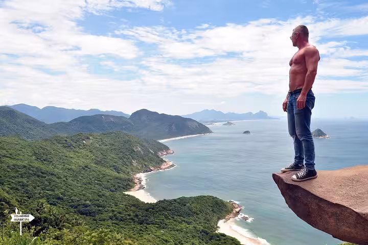 Man standing confidently on Pedra do Telégrafo rock, overlooking breathtaking ocean and forest landscape.