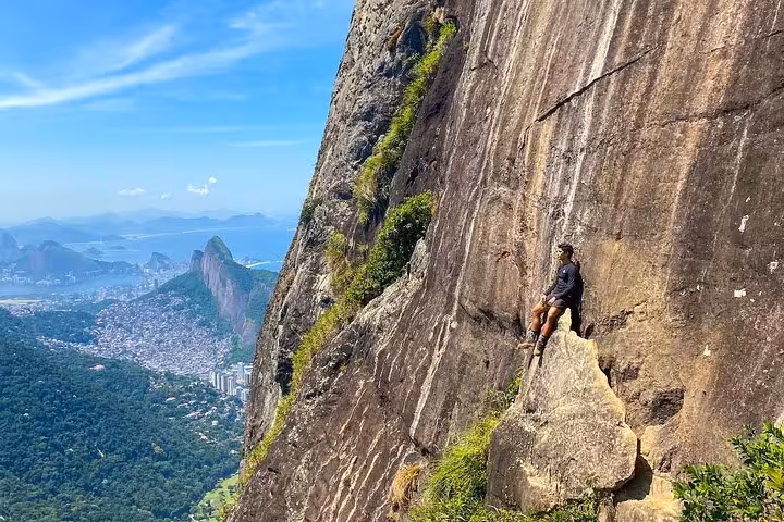 Adventurer resting on steep rock face with breathtaking view of Rio's coastline and mountains.