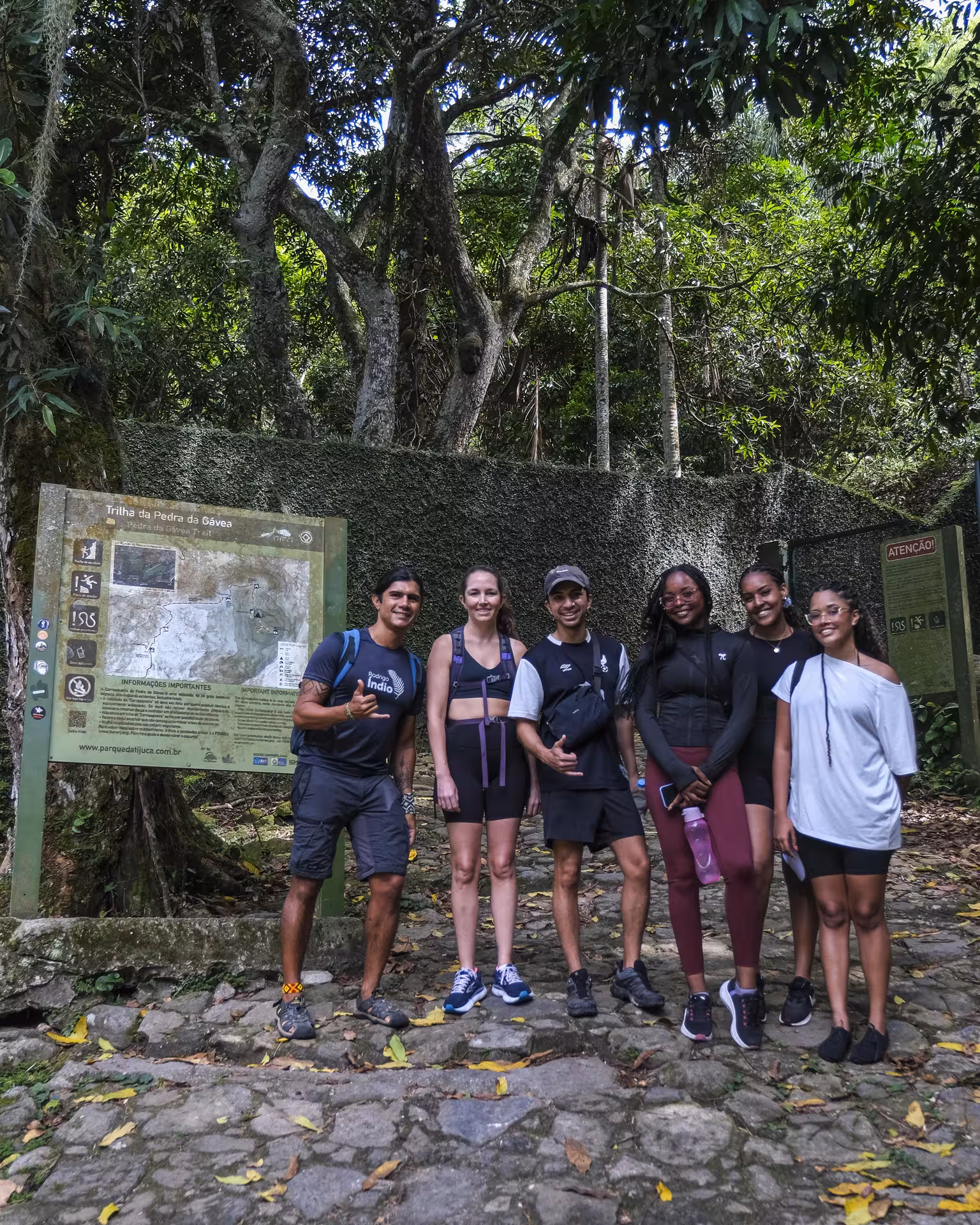 Group of hikers posing at the start of Trilha Pedra da Gávea in Rio, surrounded by lush forest and trail signs.