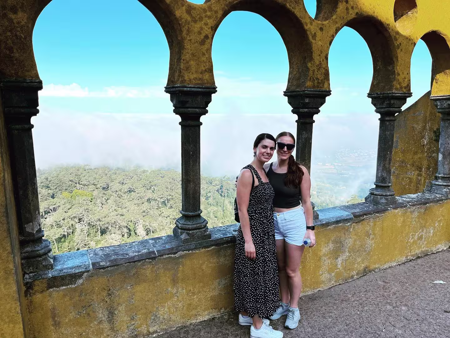 Two friends standing by the arched windows of Pena Palace, overlooking misty forested hills in Sintra.