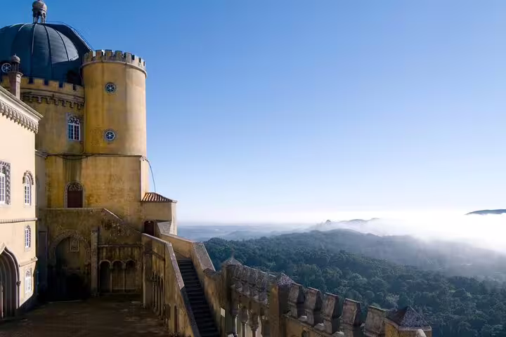 Pena Palace's vibrant architecture overlooks lush Sintra forests and misty hills on a sunny day, featured on a full-day tour.